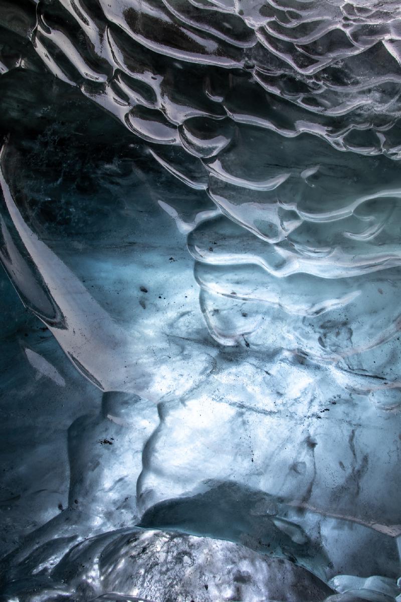 Close-up of the underside of glacier ice, its surface carved into smooth blue-grey ridges and folds, lit from within