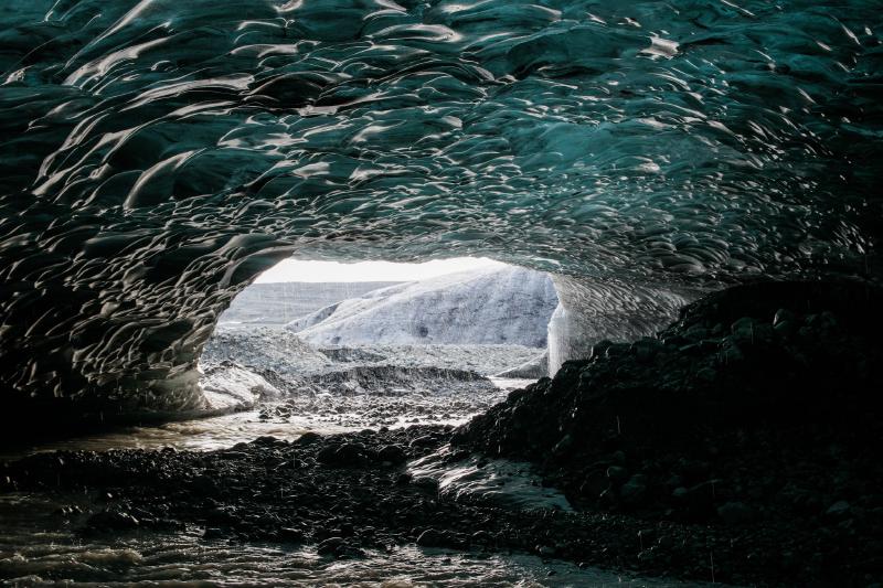 Looking out through the mouth of an ice cave, over a rocky floor and meltwater stream, toward daylight and the glacier beyond