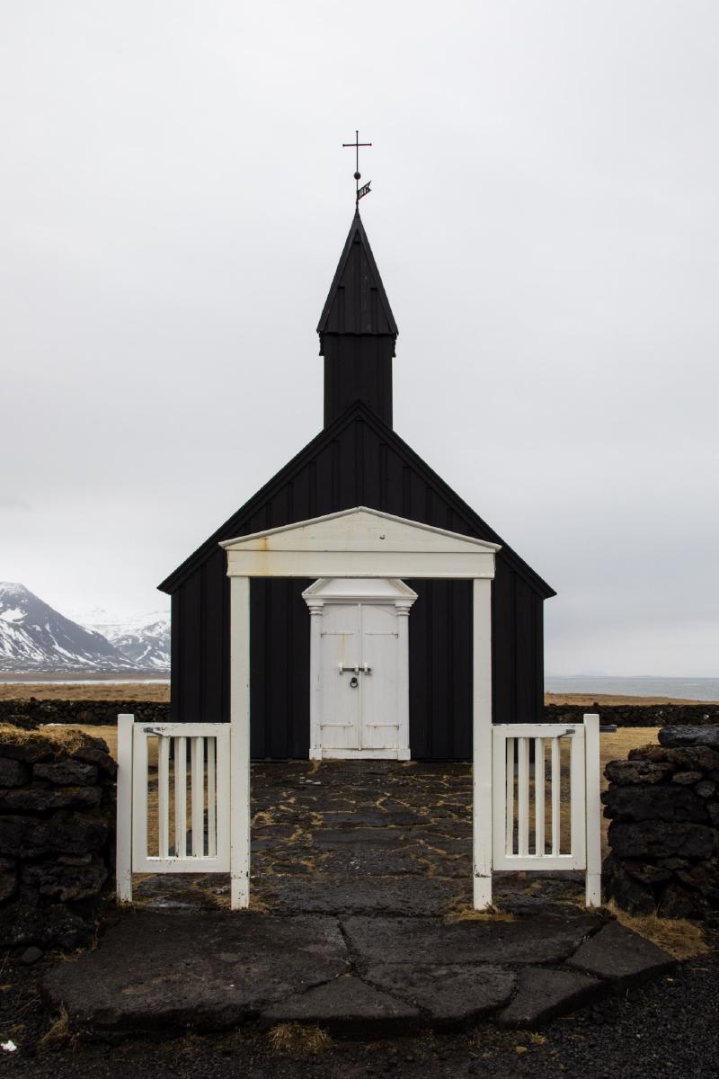 A small black timber church with a white door and gate, framed by a low lava-rock wall, snow-capped mountains behind it under an overcast sky