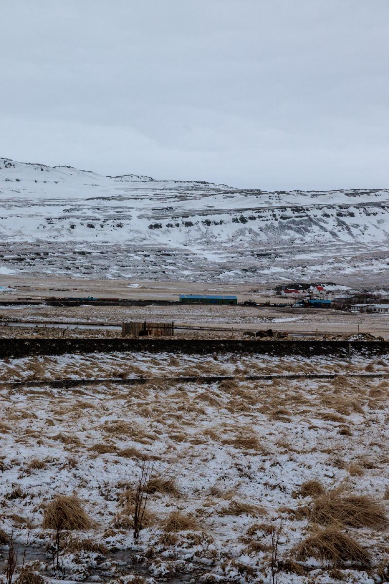 A snow-dusted valley with farm buildings at the foot of streaked, snow-covered mountains