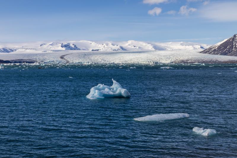 Icebergs drifting in blue water in front of a glacier and snow-covered mountains under a clear sky